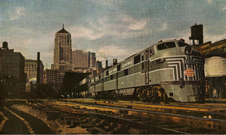 Painting of a train outside on a sunny day, in front of a city.